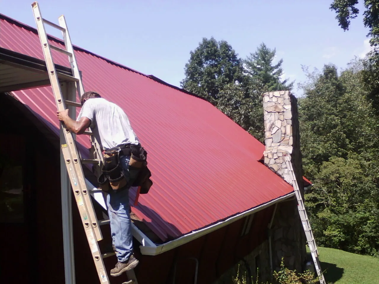 Roofer on a ladder beside a red metal roof and stone chimney — Soffit & Fascia Repair by Roofing Brothers in Barnhart